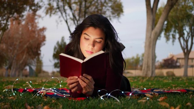A young adult woman reading a book and using her imagination to bring the story to life outdoors in the park at sunset.