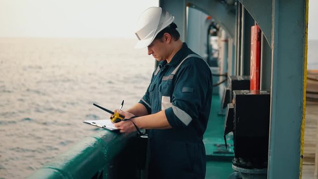 Marine chief officer or chief mate on deck of ship or vessel. He fills up ahts vessel checklist. Ship routine paperwork. He holds VHF walkie-talkie radio in hands.
