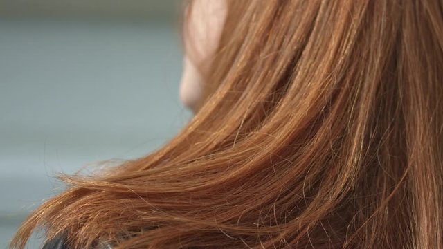 Red haired girl swaying her healthy, lush, ginger hair in the wind. Wearing a black leather jacket with a grey background.
