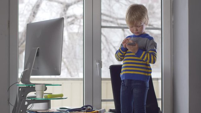 Cute blond boy is playing with smartphone, standing on his father work chair next to modern computer. child wants to quickly become adult and learn everything. Home education concept for lagging kids