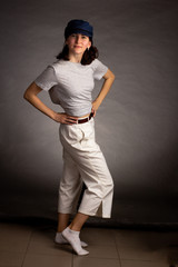 a young woman in a light t-shirt, trousers and blue cap poses on a dark background