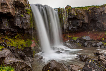 Obraz premium Waterfall falls from a small height from cliffs with a reddish tint and covered with green grass and moss.