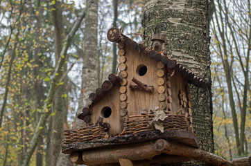 Nice wooden birdhouse attached to birch trunk made of boards and twigs with double level roof, feeder-balcony, braided twigs fence on blurred background of forest. Concept: care of wild animals