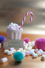 Happy Holidays handwriting on a cardboard and a cup full of tiny marshmallows, candy cane, colorful pom poms