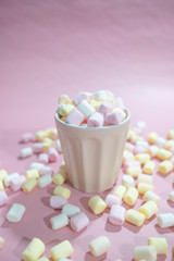 Cup full of tiny colored marshmallows and a lot of marshmallows around it on a pink background