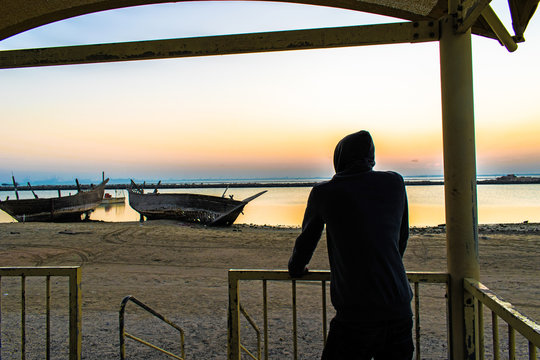 a man wearing hoodie is standing at a beach front of an abandoned broken wooden fishing boats on the side of the beach and looking so sad