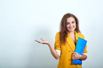 Young woman in yellow jacket keeping blue folder, pointing aside, looking happy isolated on gray...