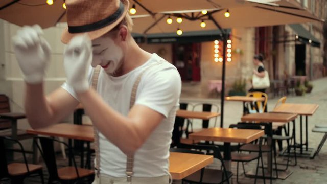 Handsome young mime in hat, suspenders and white gloves performs near street cafe. Entertainer is jumping forward, happy. Then hesitates to move further, concentrated, jumps, readjusts hat.