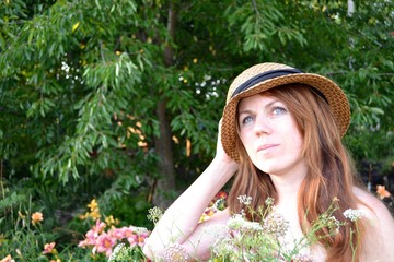 Portrait of a young woman with long brown hair in a hat and a raspberry sundress with a bouquet of wild flowers in a flowered summer garden