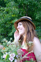 Fototapeta premium Portrait of a young woman with long brown hair in a hat and a raspberry sundress with a bouquet of wild flowers in a flowered summer garden