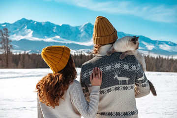 Young couple with cat in sweaters and knitted hats against mountains
