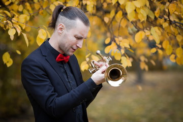 young handsome European musician in classic black suit and red bow with trumpet in autumnal park