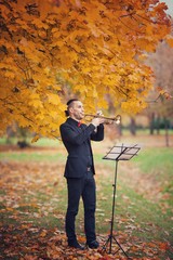 young slender handsome trumpeter blowing his trumpet near music stand with notes in autumn park...