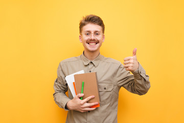 Portrait of a cheerful nerd in a shirt and glasses stands on a yellow background with books and exercise books in his hands, looks in camera, shows his thumb up and smiles. Learning concept. Isolated