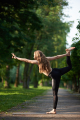 Woman with long hair in black sports suit standing in pose of king of dance, Asan Natarajasana, against greenery park under sunny rays. Morning training outdoors at summer. Healthy lifestyle concept