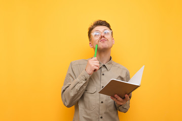 Portrait of pensive student on yellow background with exercise book and pen in hand on yellow...