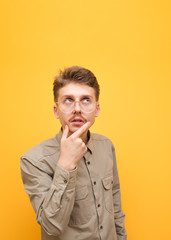 Portrait of funny nerd student on yellow background, wears glasses and beige shirt, looks up at copy space and thinks. Guy geek with mustache and glasses pensive, isolated on yellow background.
