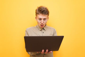 Surprised nerd in glasses and shirt works on laptop on yellow background, with shocked face looks into screen. Emotional freelancer stands with a laptop in his hands on a yellow background © bodnarphoto