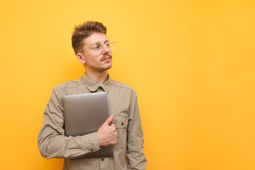 Funny student with a laptop in his hand stands on a yellow background and looks away at copy space,...
