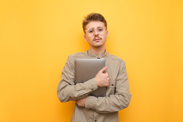 Portrait of serious fashion man in glasses and shirt standing with laptop in hand on yellow background, looking into camera with serious face. Programmer with laptop posing on camera. Isolated.