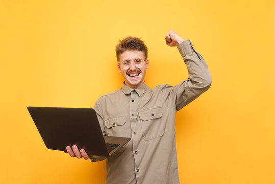 Portrait of joyful programmer with laptop in hand on yellow background, rejoices in victory and looks at camera, wears glasses and shirt. Happy guy in mustache and glasses rejoices. Isolated.