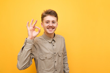 Happy guy with mustache and glasses shows OK gesture and smiles against yellow background, looks into camera and wears shirt. Cheerful student shows hand gesture alright. Copy space