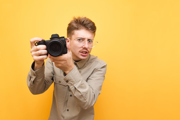 Obraz premium Portrait of funny paparazzi photographer with glasses and wears shirt on yellow background, holds camera in hand and makes photo, looks into camera with serious face. Copy space