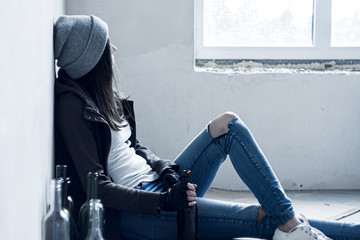 Homeless drunk young woman in hat is sitting on floor in abandoned building. Addicted teenage girl...