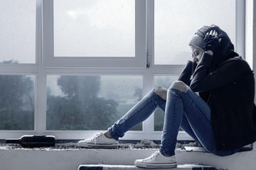 Lonely, depressed, upset young woman in hood is sitting on windowsill and crying in abandoned building. Homeless teenage girl ran away from home because of problems. Alcohol abuse, addiction concept. - obrazy, fototapety, plakaty