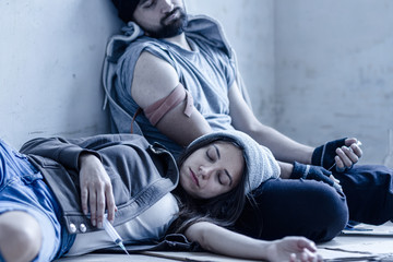 Homeless young man and woman are lying on cardboard on floor in abandoned house. Guy and girl addicts got overdose injections of heroin and getting high. Drug addiction and street life concept. - obrazy, fototapety, plakaty