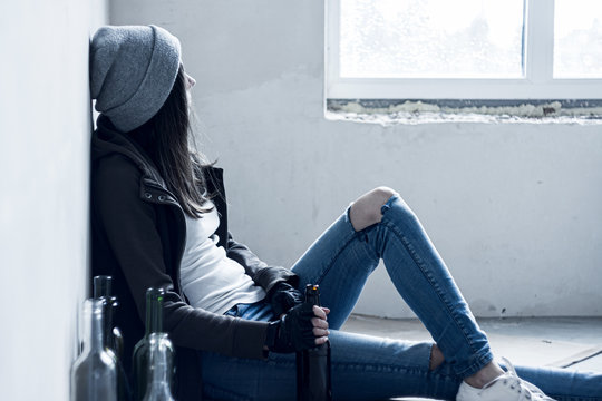 Homeless drunk young woman in hat is sitting on floor in abandoned building. Addicted teenage girl drunkard is drinking beer. Bum is living in street. Alcoholism, alcohol abuse concept.