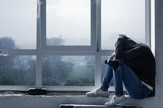 Lonely, depressed, upset young woman in hood is sitting on windowsill and crying in abandoned building. Homeless teenage girl ran away from home because of problems. Alcohol abuse, addiction concept.