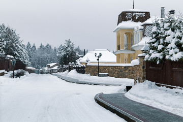 Snowy road between country houses. Winter landscape road and brick sidewalk (pavement). Street landscape.