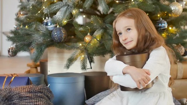 Smiling cute blond girl is hugging a gold gift box she recieved sitting near christmas tree, happy childhood and xmas celebration concepts