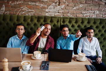 Group of four south asian men's posed at business meeting in cafe. Indians work with laptops together using various gadgets, having conversation. Hold credit card to pay.