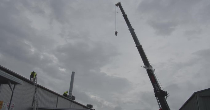 A hydraulic mobile crane with tradies wearing hi-viz shirts atop an industrial shed roof