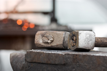 Old rusty hammer lies on the anvil with flame of brazier forge on background. Blacksmith, metalsmith, farrier tools. Close view.