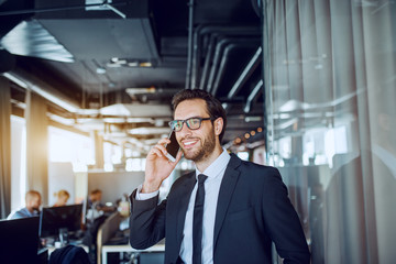 Young attractive Caucasian bearded smiling businessman in suit and with eyeglasses standing in...