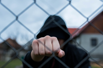 Aggressive  teenage boy showing hes fist behind wired fence at the correctional institute,  focus...