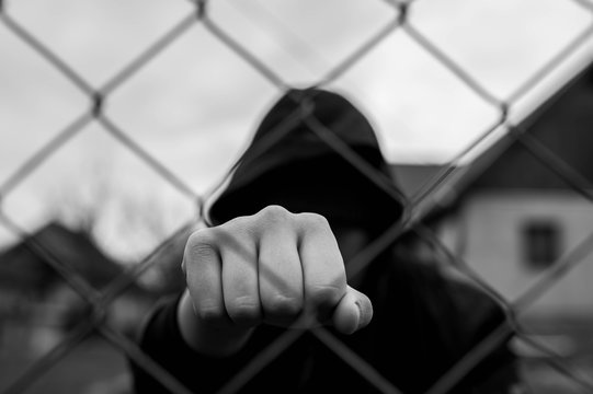 Aggressive  teenage boy showing hes fist behind wired fence at the correctional institute,  focus on the boys hand , conceptual image of juvenile delinquency in black and white.