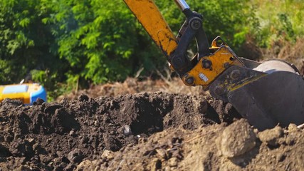 Bucket of tractor digs the ground. Excavator digging the soil on the background of green trees in summer. Close-up.