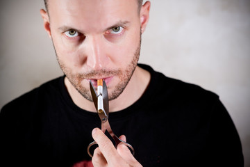 a man with a three-day stubble holds a cigarette in his mouth with scissors