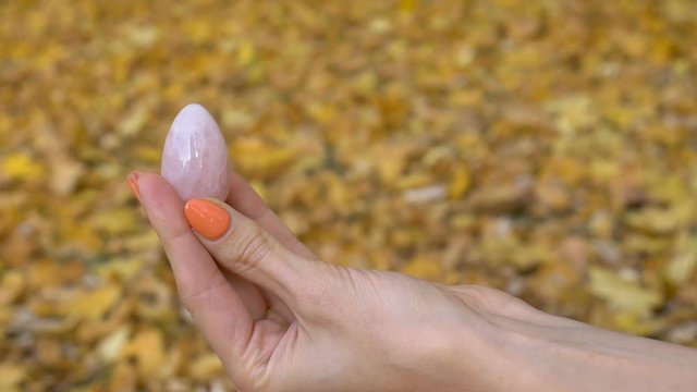 Female hand with orange manicure holding pink quartz yoni egg for vumfit, imbuilding or meditation on yellow fallen leaves background during autumn day outdoors