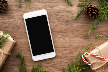 Smartphone with blank screen is in the middle of wood table with christmas decorations. Top view, flat lay.