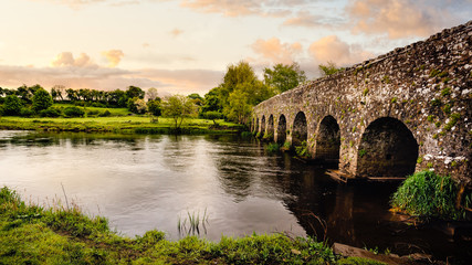Old 12th century stone arch bridge over a river. Green fields and trees. Dramatic sky sunset. Count Meath, Ireland