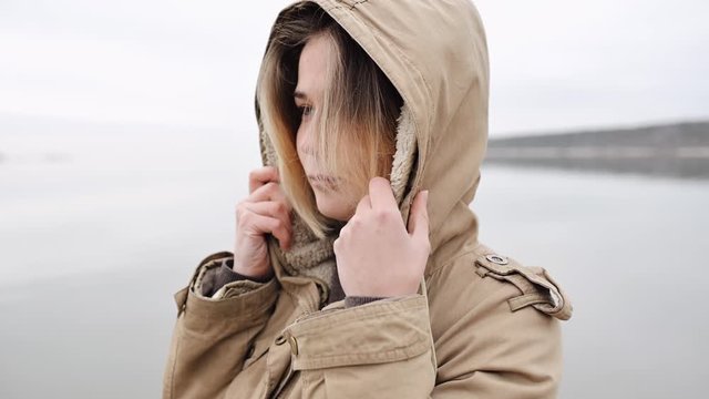 portrait of a young calm and serene girl with short blond hair standing on the background of the sea, looking into the distance and putting on the hood of a warm jacket