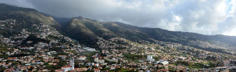 Fototapeta na wymiar Wide panoramic view of Madeira island. Town with buildings on a steep mountain. Cloudy skies in background