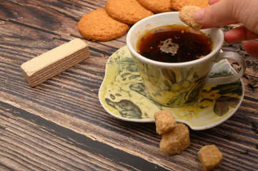 The girl's hand puts a piece of brown sugar in a mug of black tea, oatmeal cookies, tea leaves, waffles, brown sugar on a wooden background. Close up.