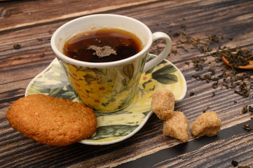 A Cup of black tea, tea leaves, pieces of brown sugar, oatmeal cookies on a wooden background. Close up.