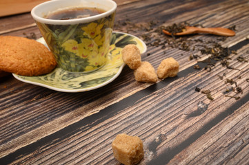 A Cup of black tea, tea leaves, pieces of brown sugar, oatmeal cookies on a wooden background. Close up.